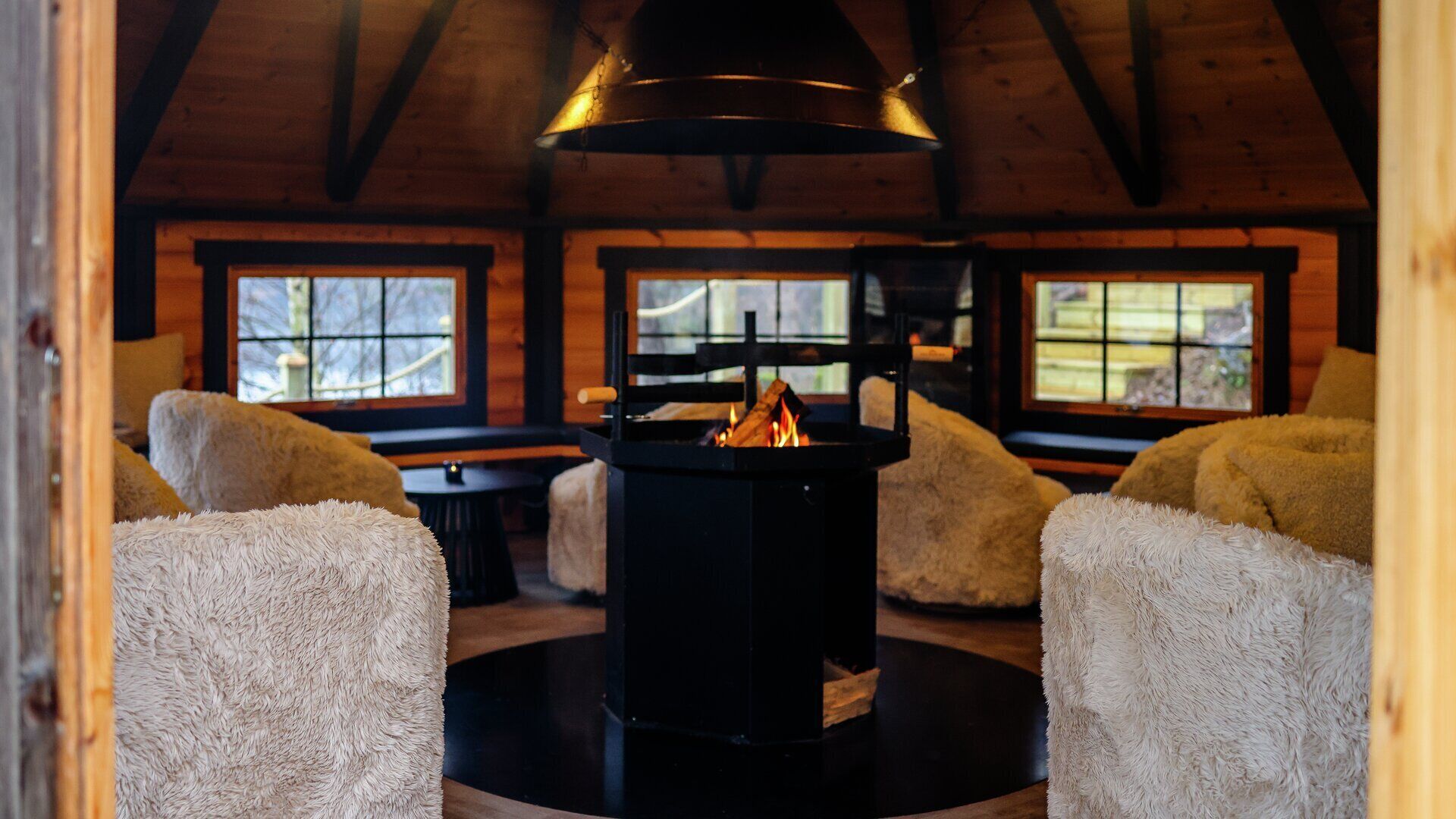 A cozy cabin interior with a central wood-burning stove, plush fur-covered chairs, and large windows offering views of the outdoors.