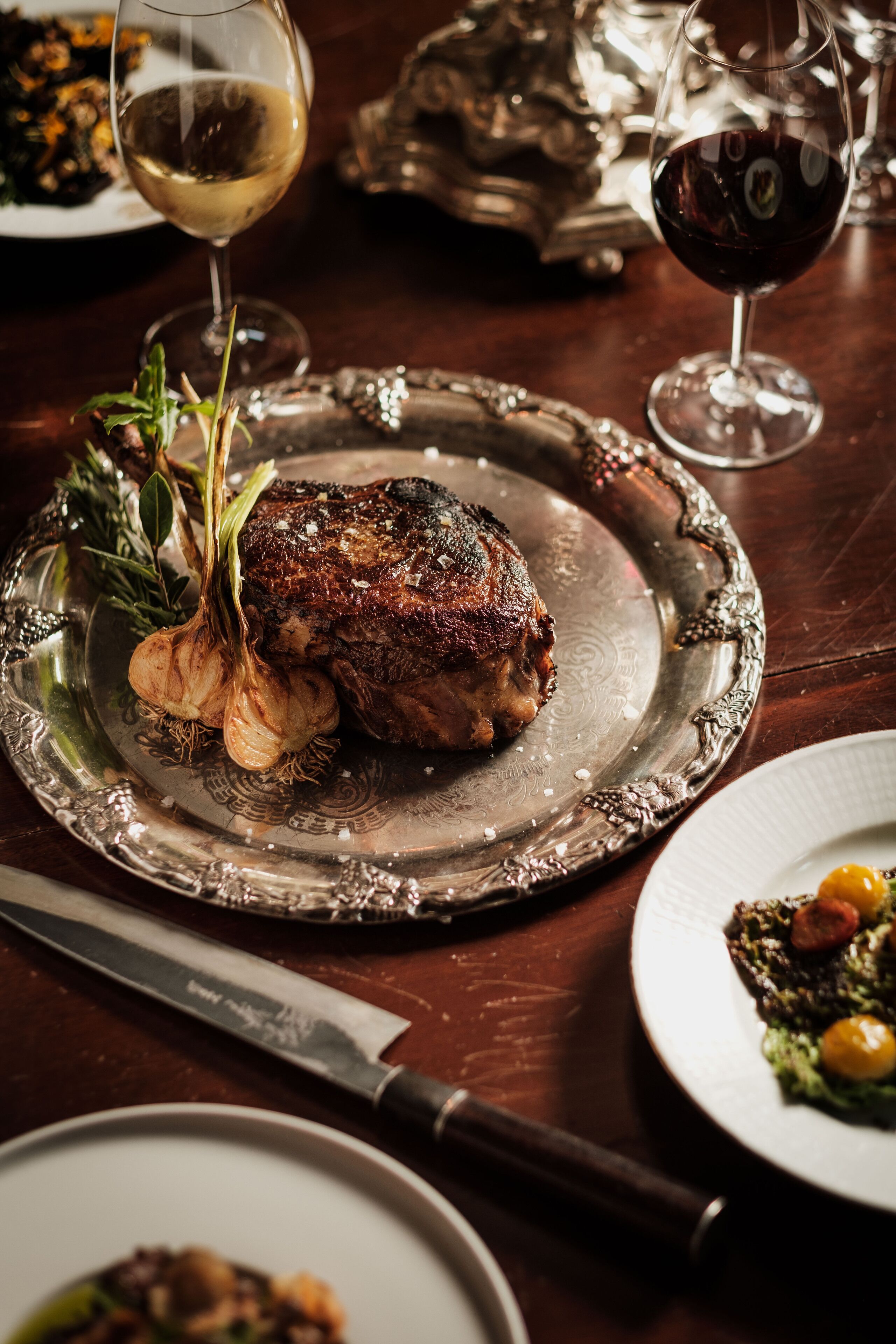 A steak with garlic and herbs on a silver plate, accompanied by wine glasses and a knife on a wooden table.