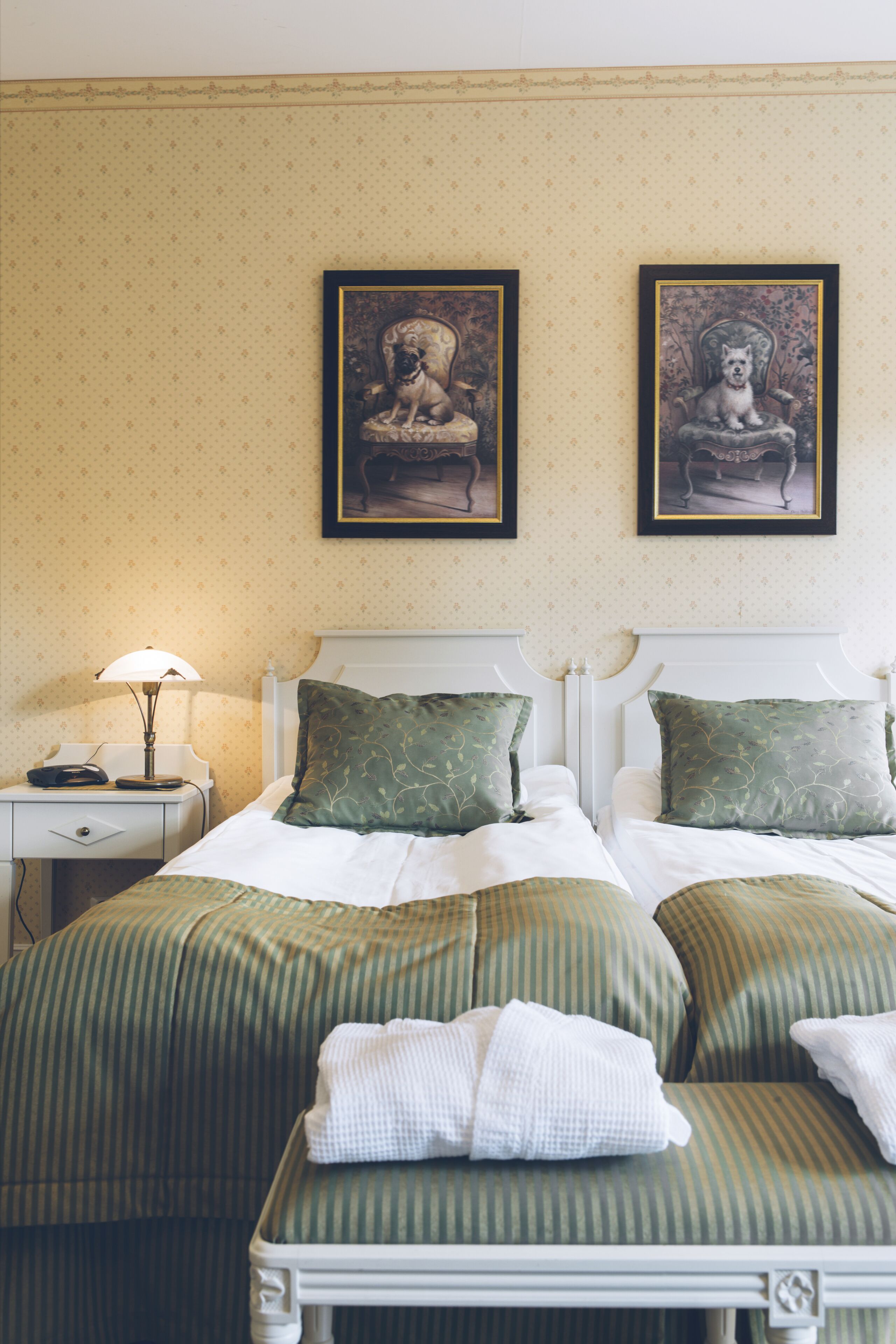 A hotel room with two beds, green striped bedding, a nightstand with a lamp, and two framed pictures on the wall.
