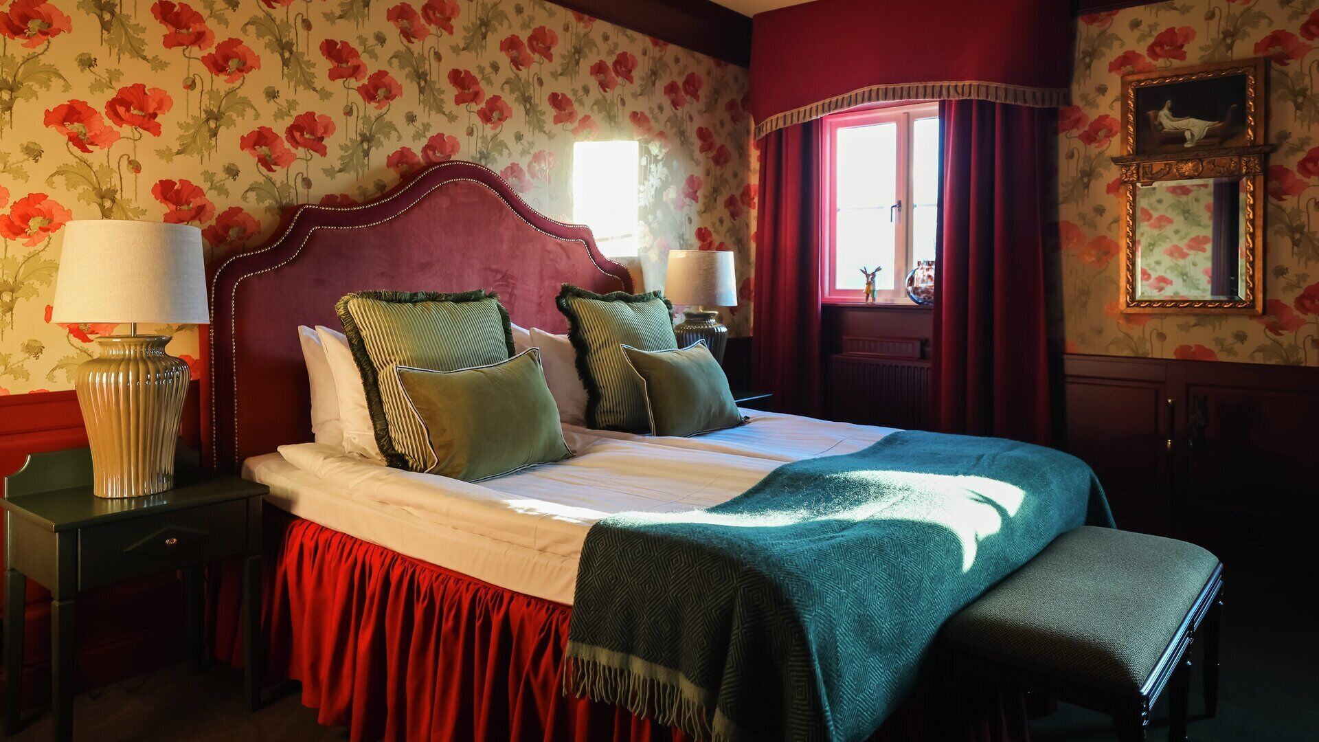 Een hotelkamer met een groot bed, twee nachtkastjes met lampen, een rood gordijn, een spiegel en bloemenbehang.