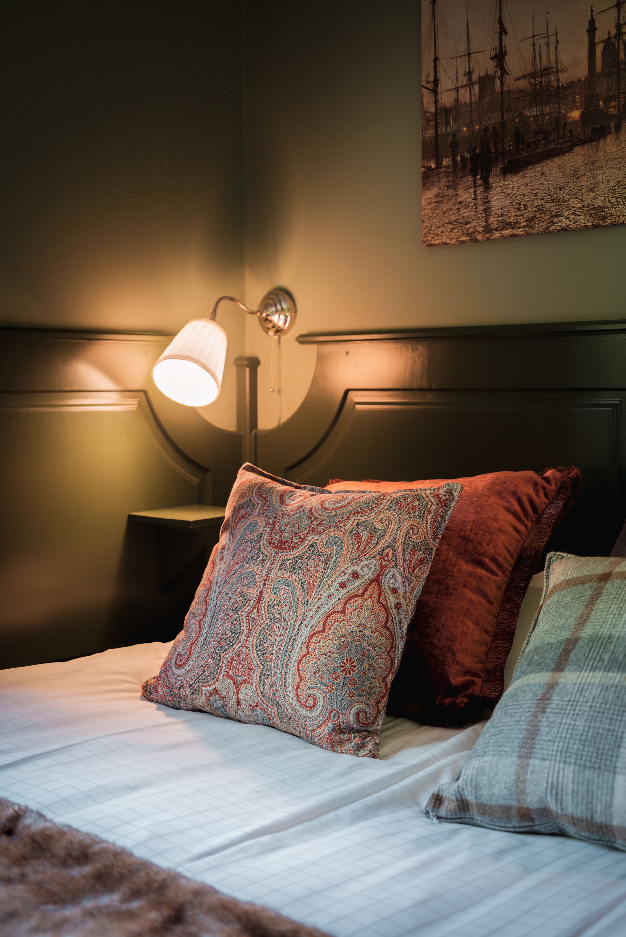 A neatly made bed with patterned pillows, a wall-mounted lamp, and a framed picture above the bed.
