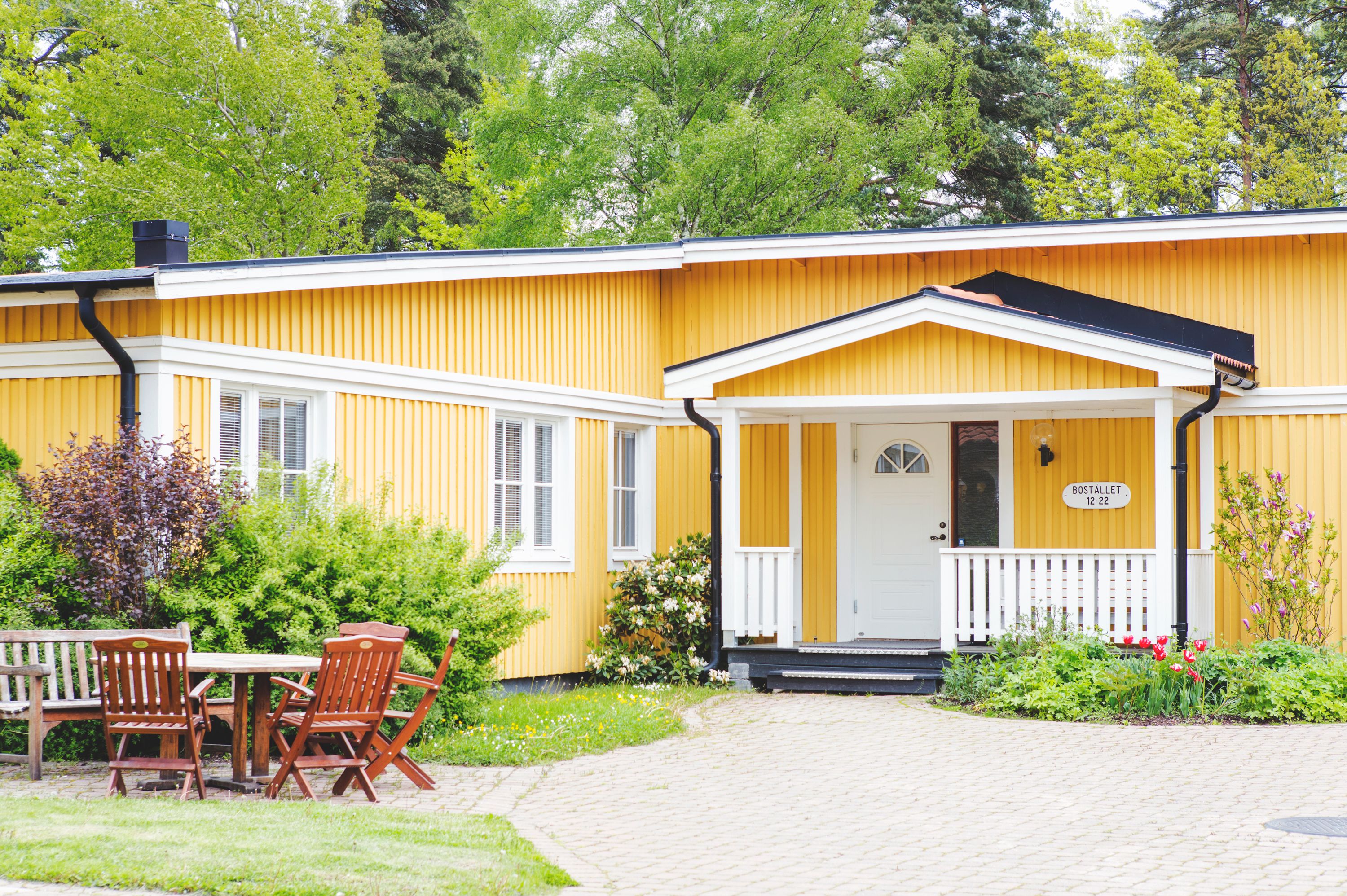 A yellow building with a white porch and a sign that reads "Bostaliet 12:22".
