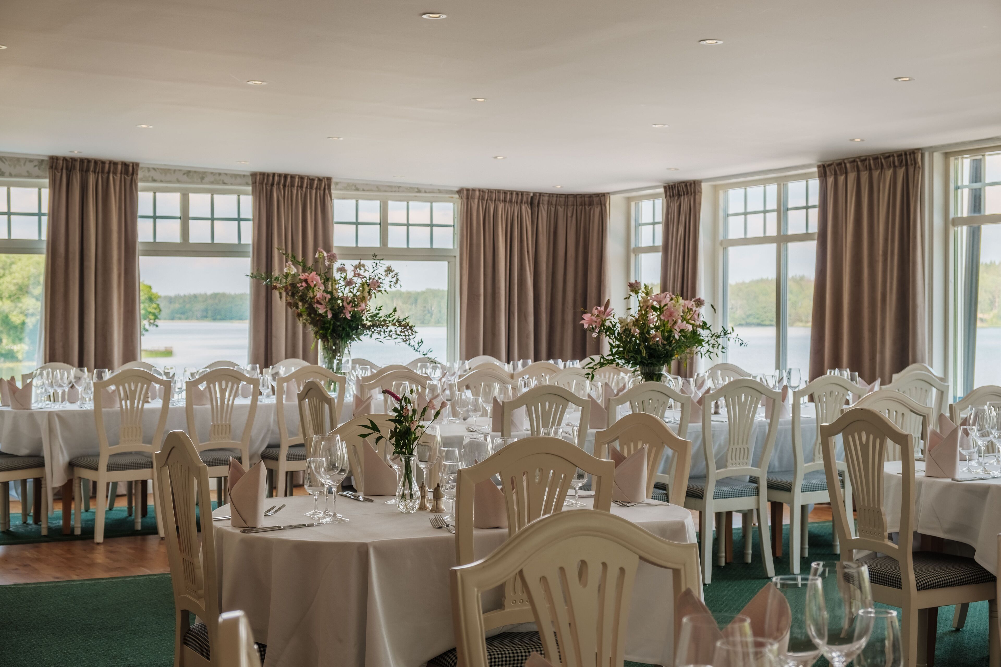 A formal dining room with multiple tables set for a meal, large windows with curtains, and a view of the outdoors.