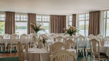 A formal dining room with multiple tables set for a meal, large windows with curtains, and a view of the outdoors.