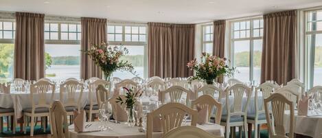 A formal dining room with multiple tables set for a meal, large windows with curtains, and a view of the outdoors.