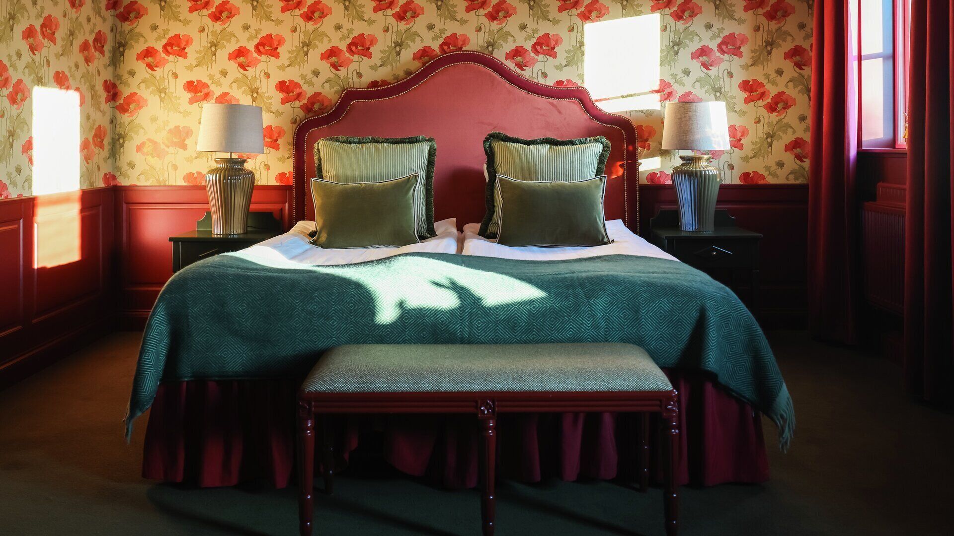 A bedroom with a large bed, two bedside lamps, a bench, and floral wallpaper.