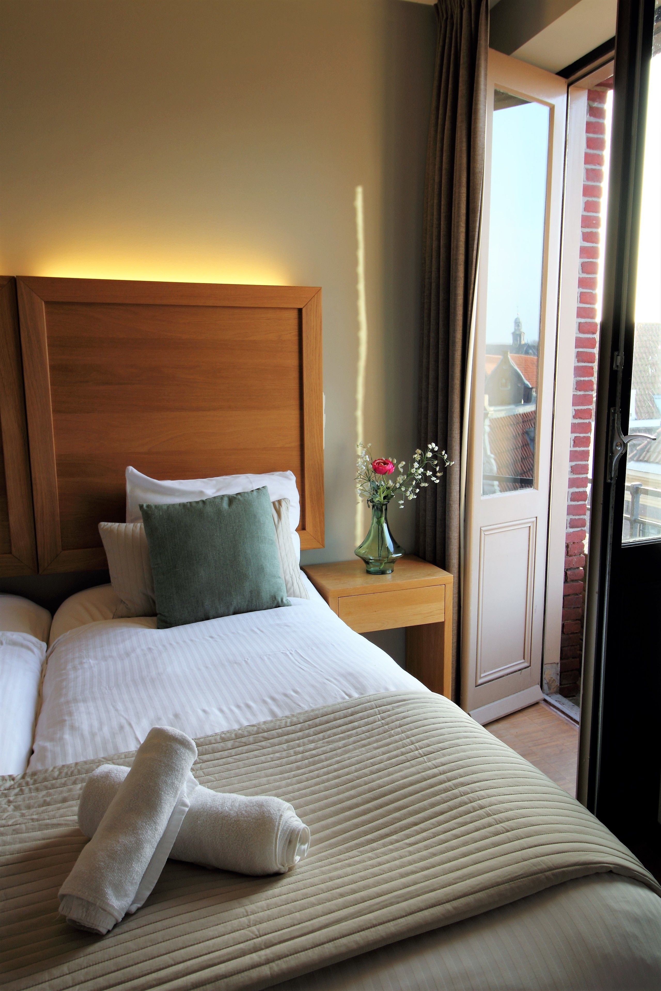 A neatly made bed with pillows, a vase with flowers, and a pair of towels on a wooden nightstand.