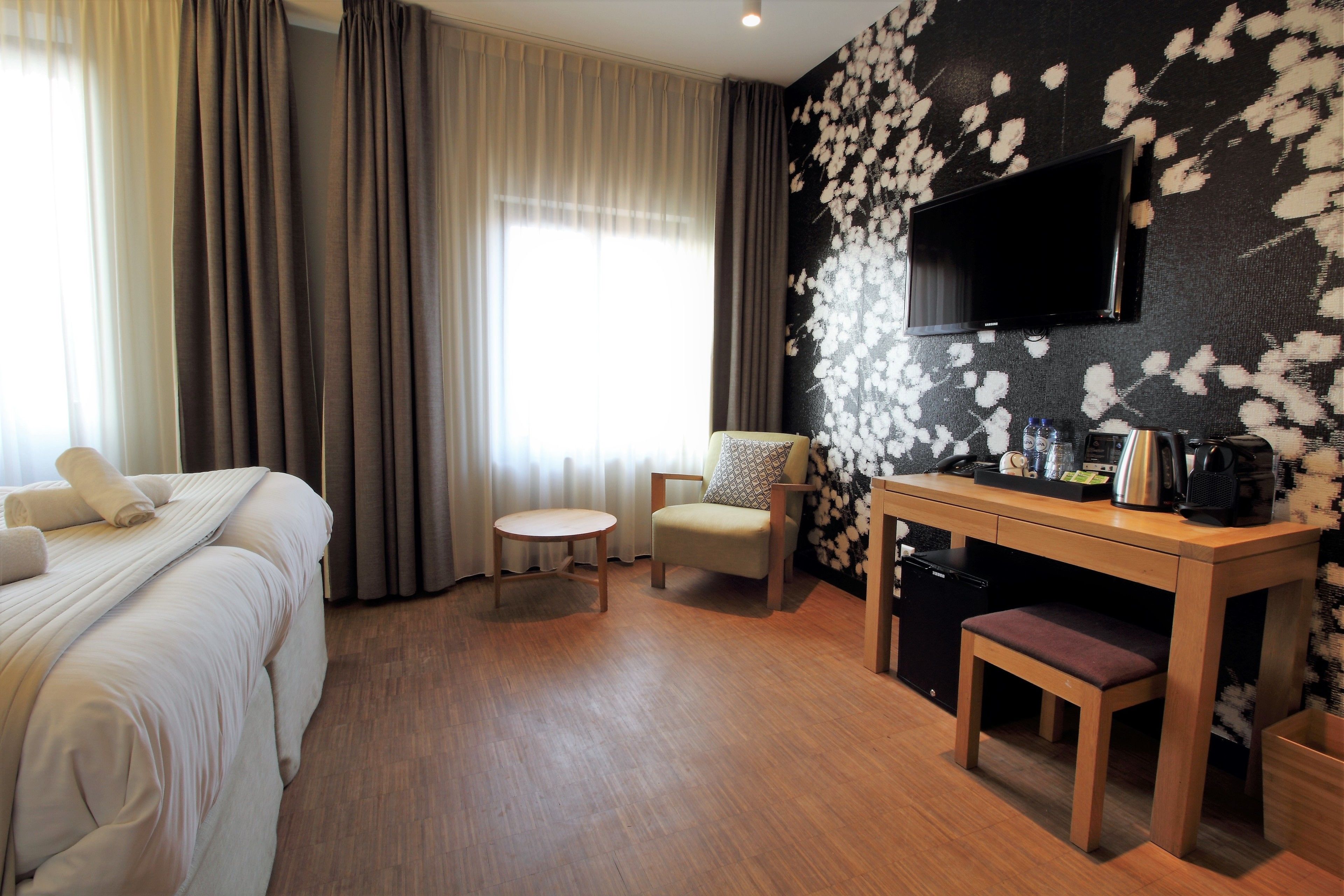 Une chambre d’hôtel comprenant un lit, un bureau, une chaise, une petite table, un téléviseur fixé au mur et une fenêtre avec des rideaux.