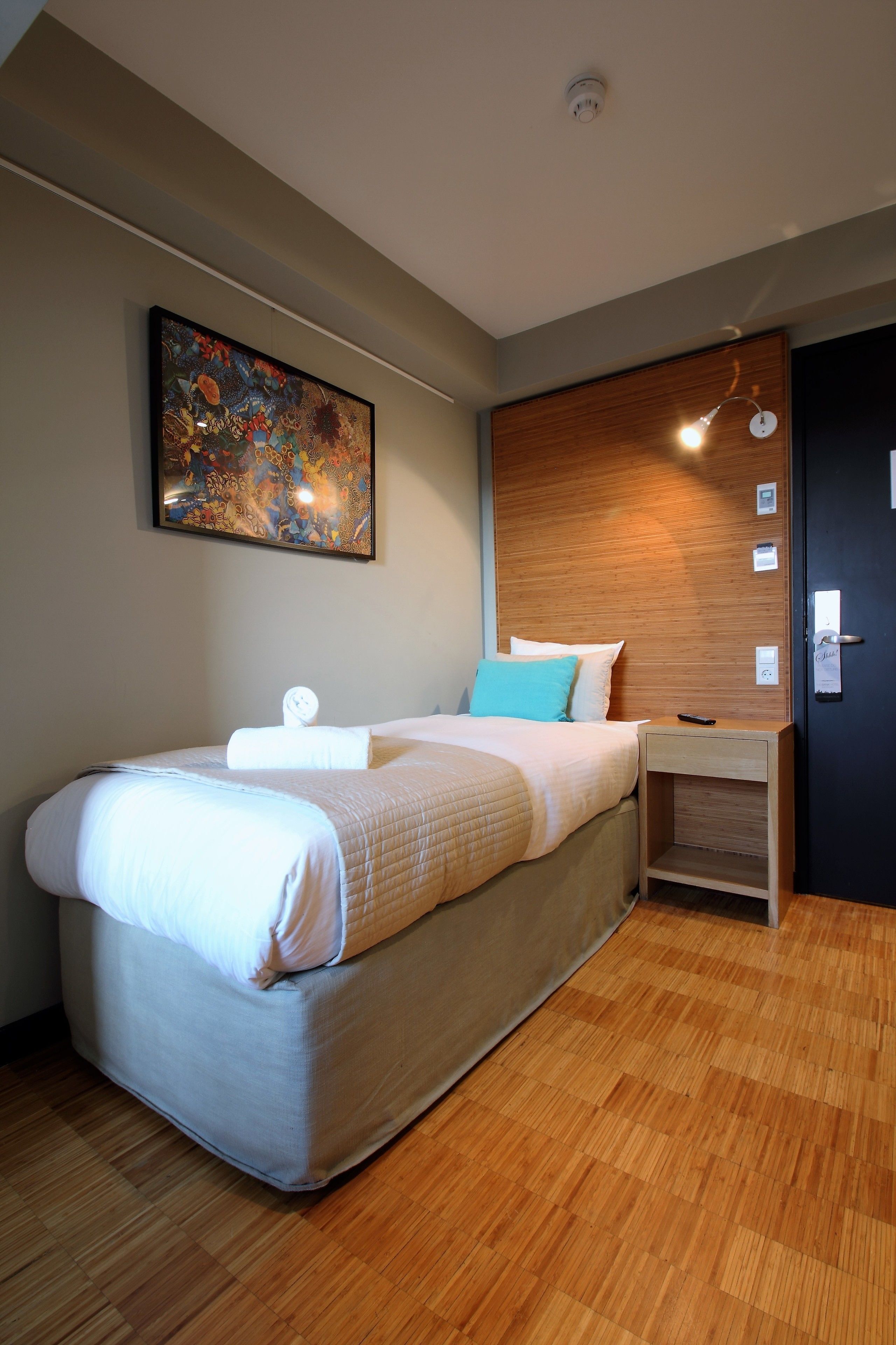 Une chambre d’hôtel moderne avec un grand lit, une tête de lit en bois, un tableau accroché au mur, une lampe de chevet et une porte dotée d’une poignée.