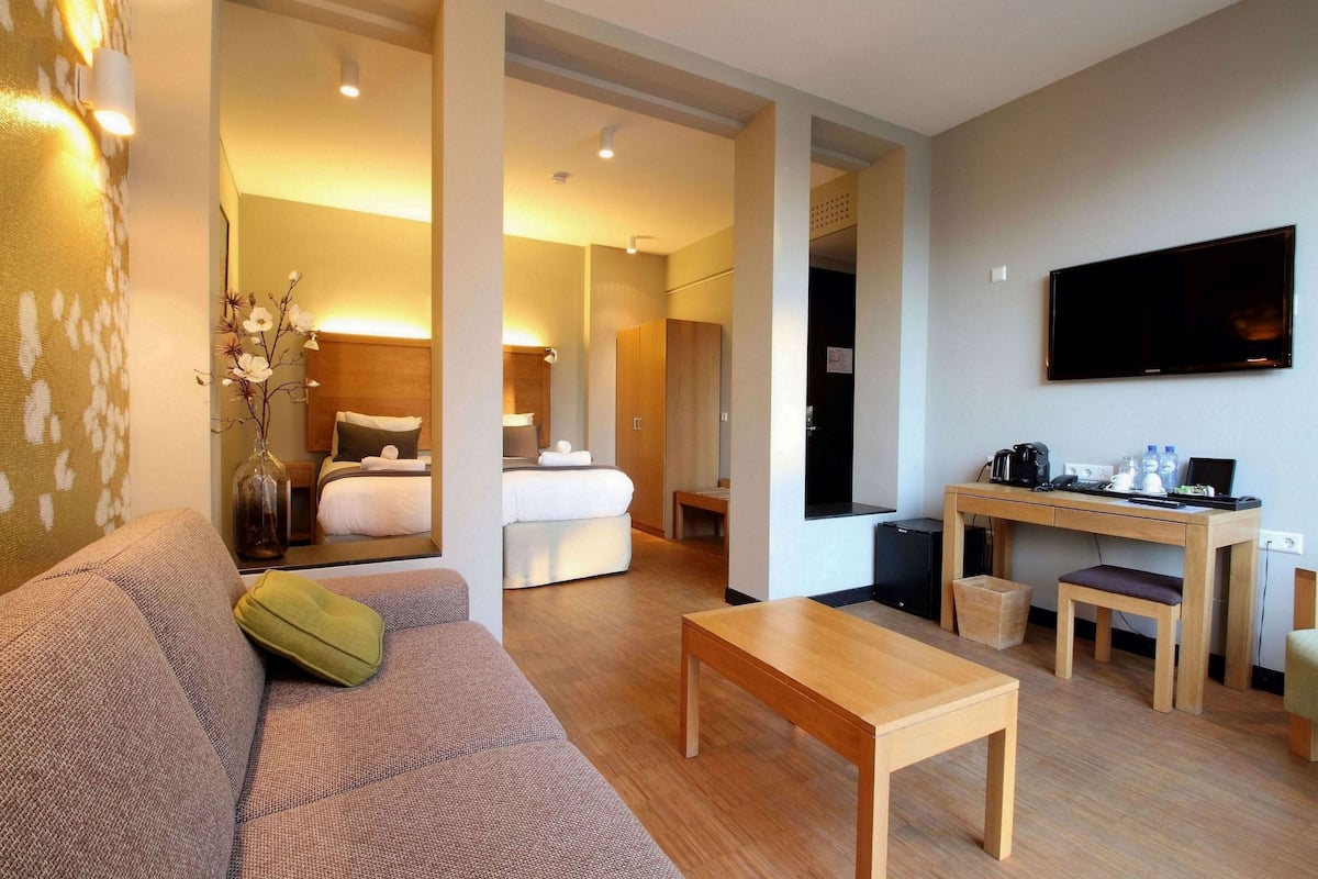 A modern hotel room with a sofa, a wooden table, a bed, a desk with a computer, and a TV.