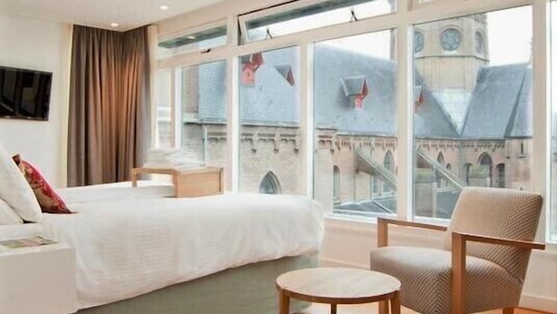 Een hotelkamer met een groot bed, een klein rond tafeltje, een stoel en uitzicht op een gebouw met een klokkentoren door grote ramen.