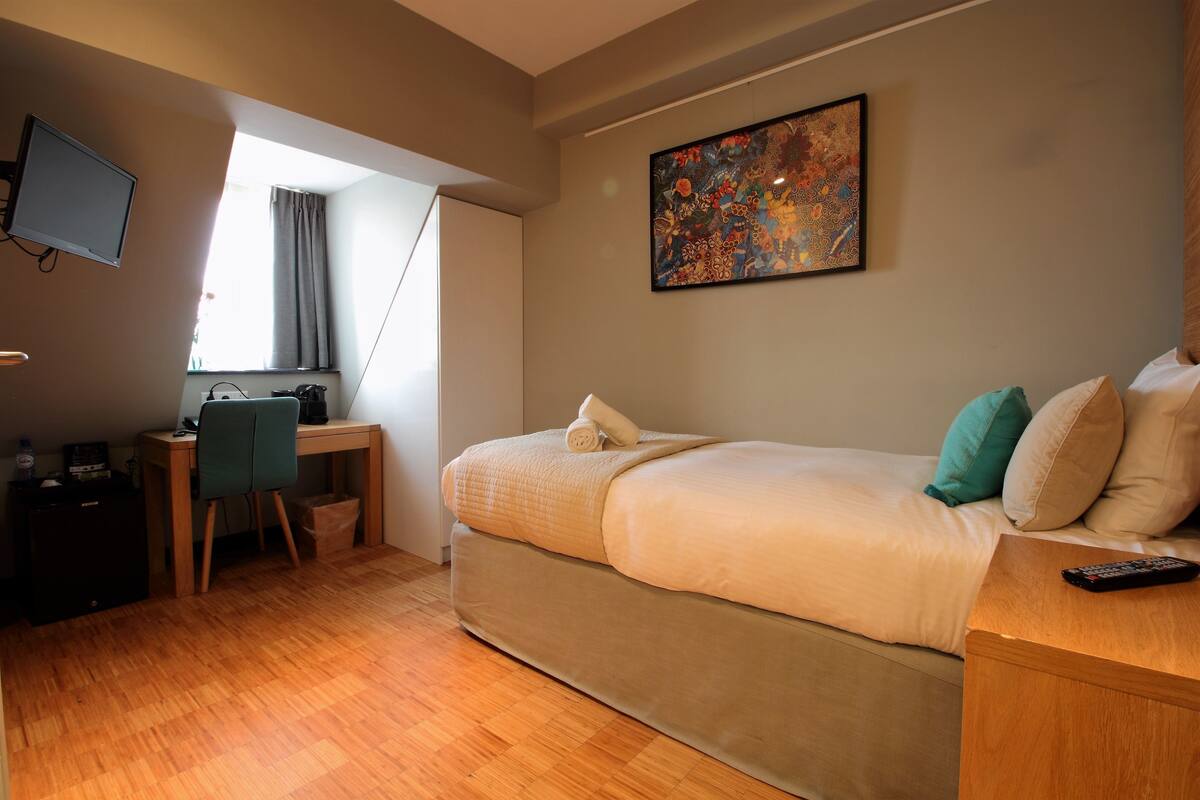 A bedroom with a bed, a desk, a chair, a TV, and a painting on the wall.