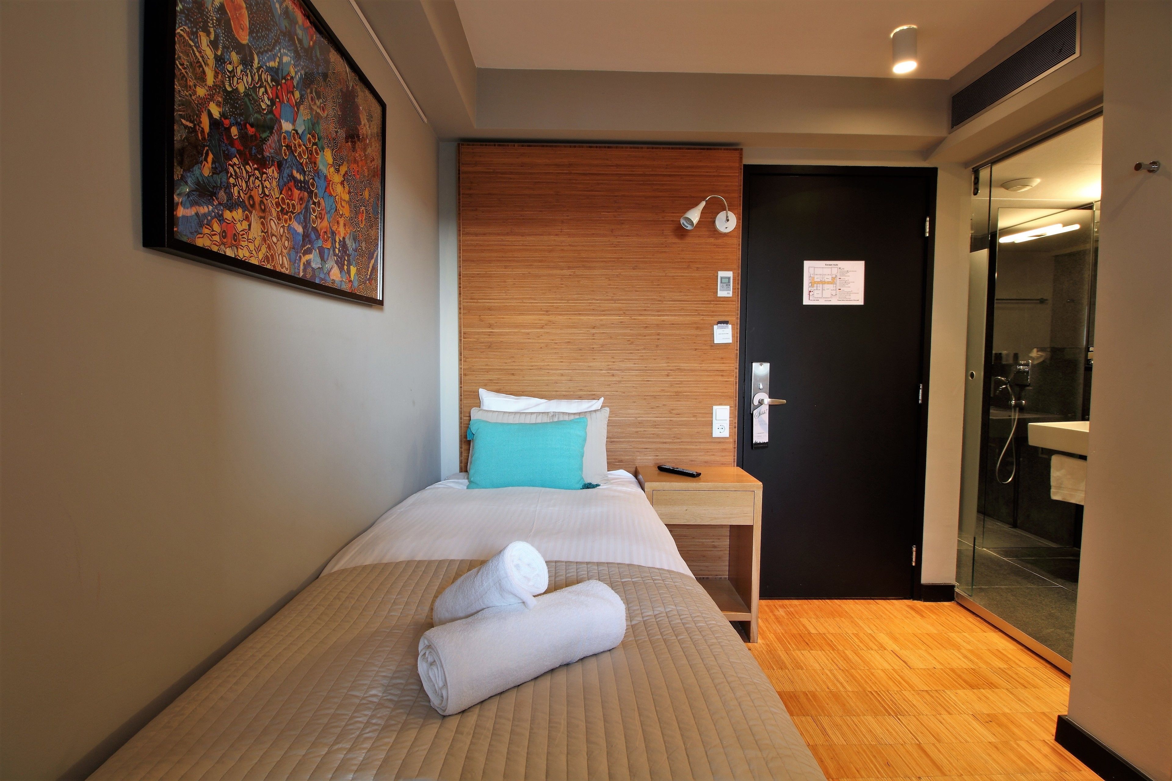 A hotel room with a bed, a wooden headboard, a painting on the wall, a nightstand, a bathroom with a glass door, and a map on the door.