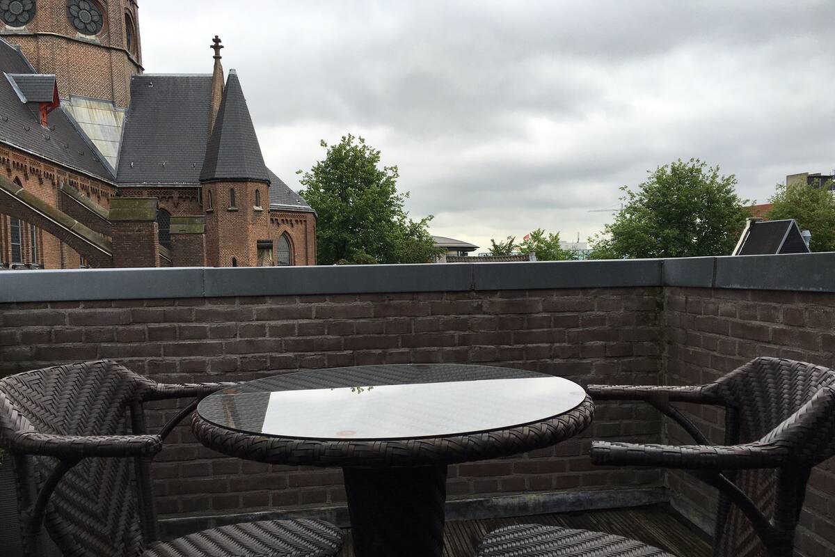 A rooftop terrace with wicker chairs and a round table, overlooking a historic building with a clock tower.