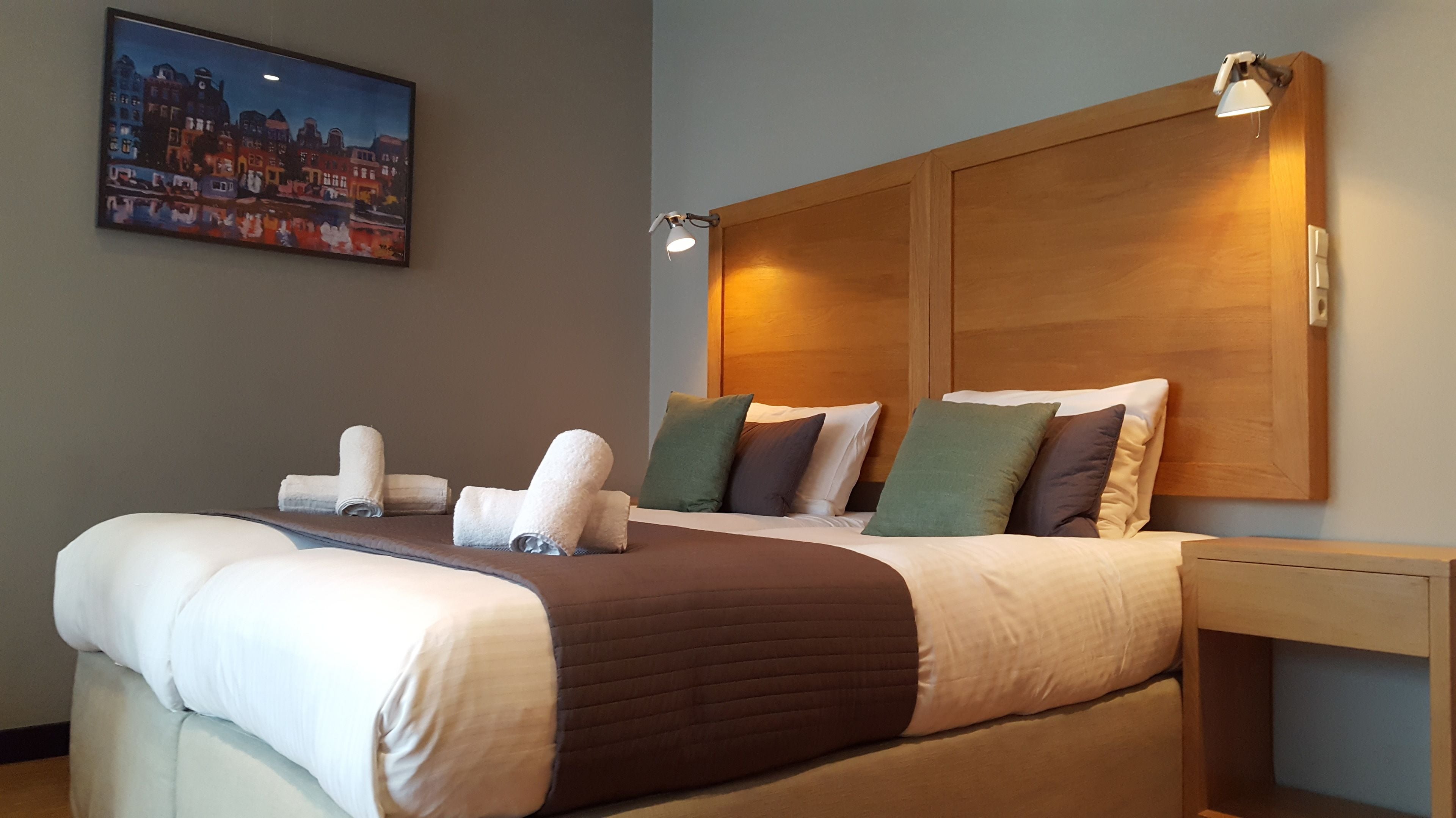 Un lit bien fait, avec une tête de lit marron, une table de chevet sur laquelle se trouve une lampe, et un tableau représentant un paysage urbain accroché au mur.