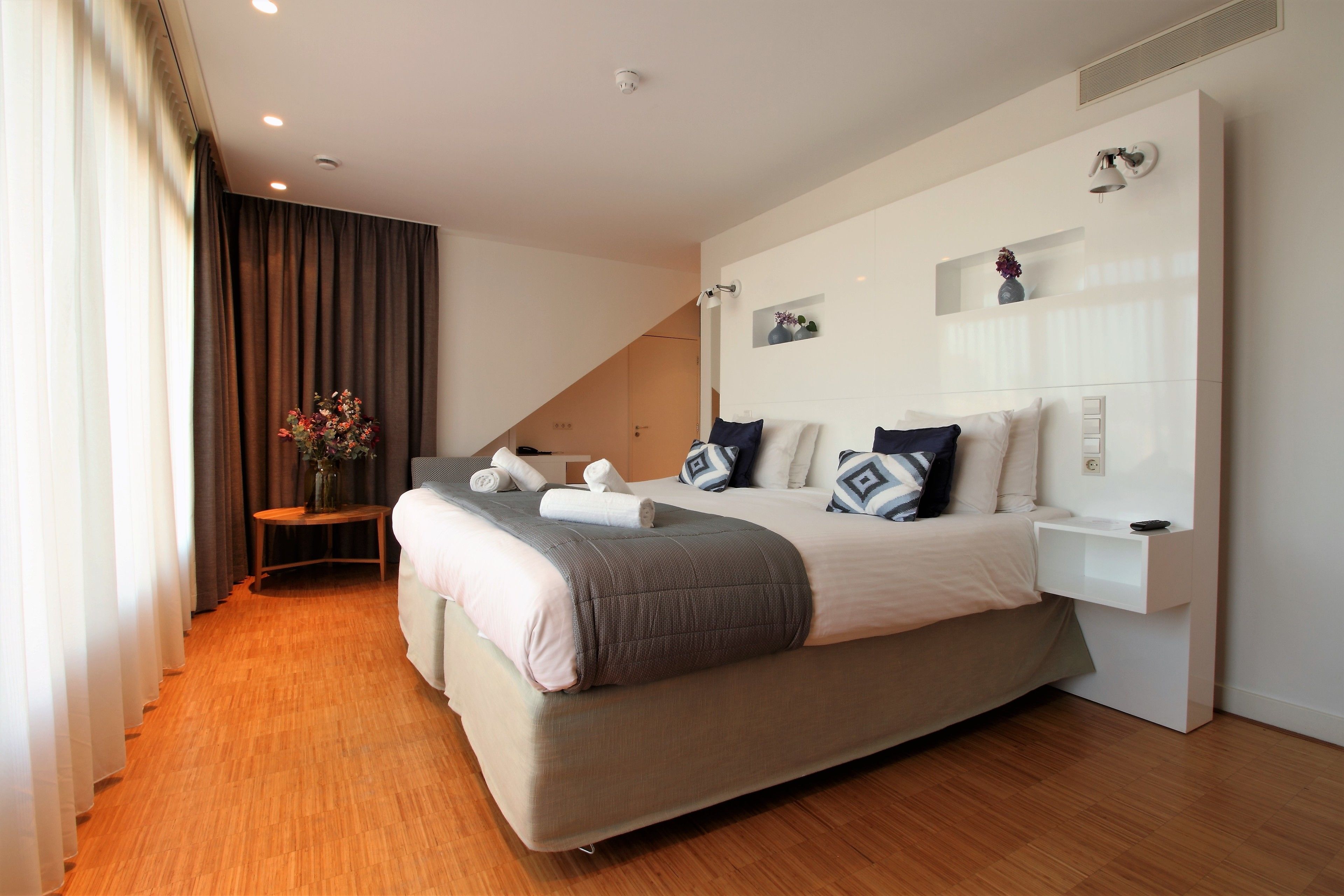 Um quarto moderno com uma cama grande, uma mesa de cabeceira, uma pequena mesa com um vaso e um prateleira fixada na parede.