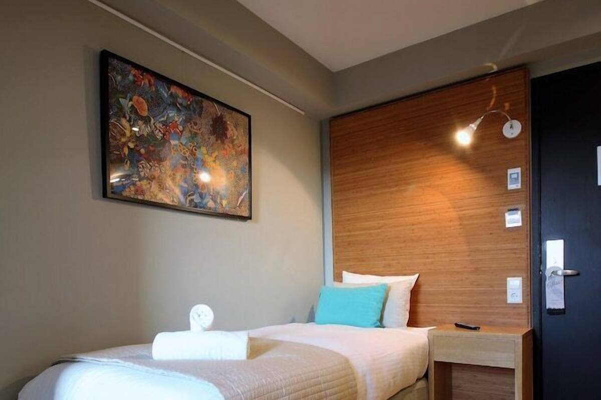 A modern hotel room with a large bed, a wooden headboard, a wall-mounted painting, a bedside table with a lamp, and a door with a handle.