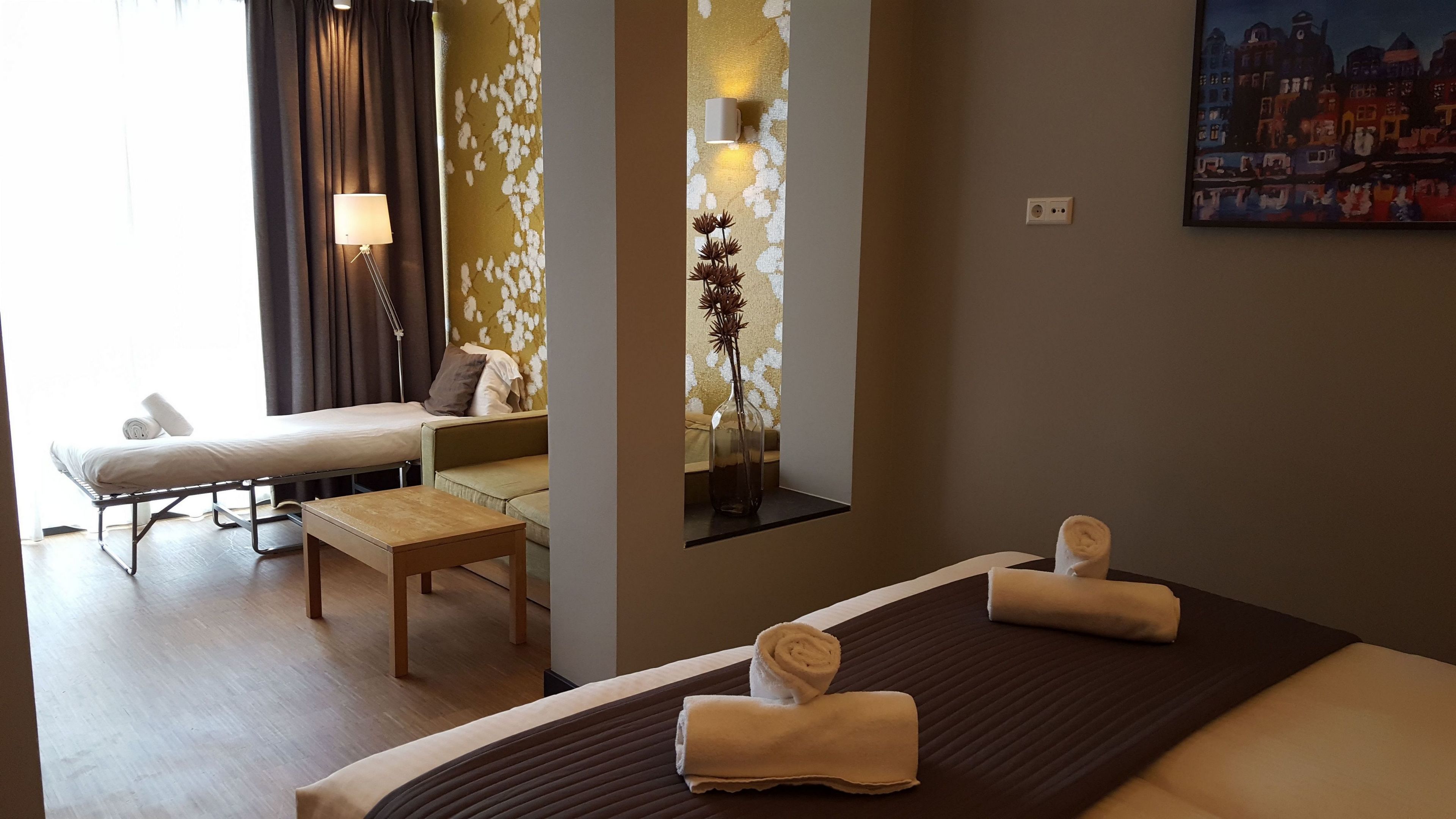 A hotel room with a bed, a sofa, a small table, a lamp, a vase with flowers, and a painting on the wall.