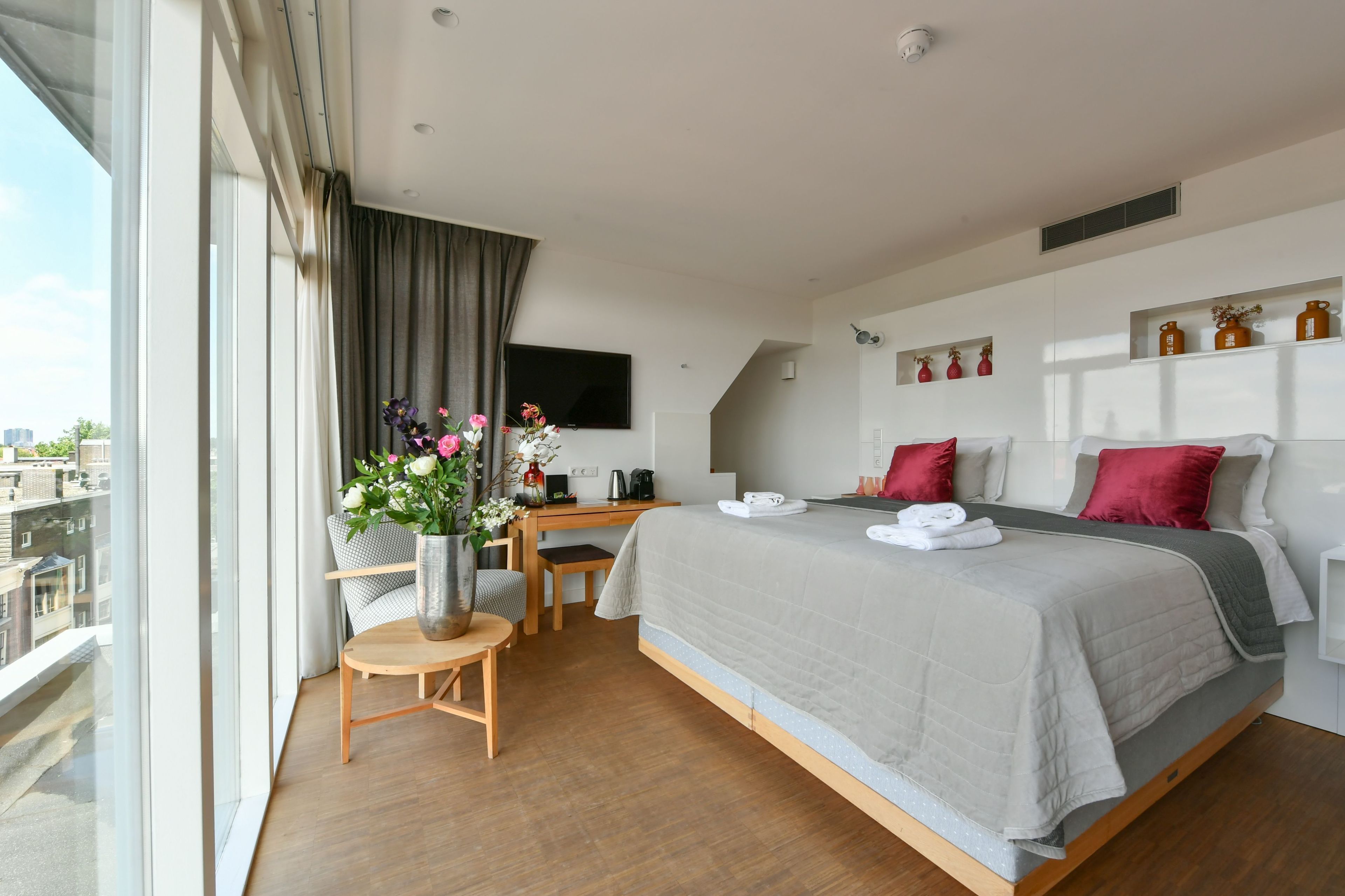 Ein modernes Hotelzimmer mit einem großen Bett, einem Schreibtisch, einem Sessel, einem kleinen Tisch, einer Vase mit Blumen und einem Fernseher an der Wand.