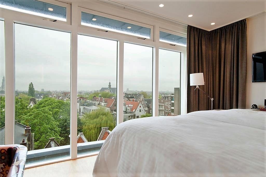A bedroom with a large bed, a view of a cityscape through floor-to-ceiling windows, and a modern lamp.