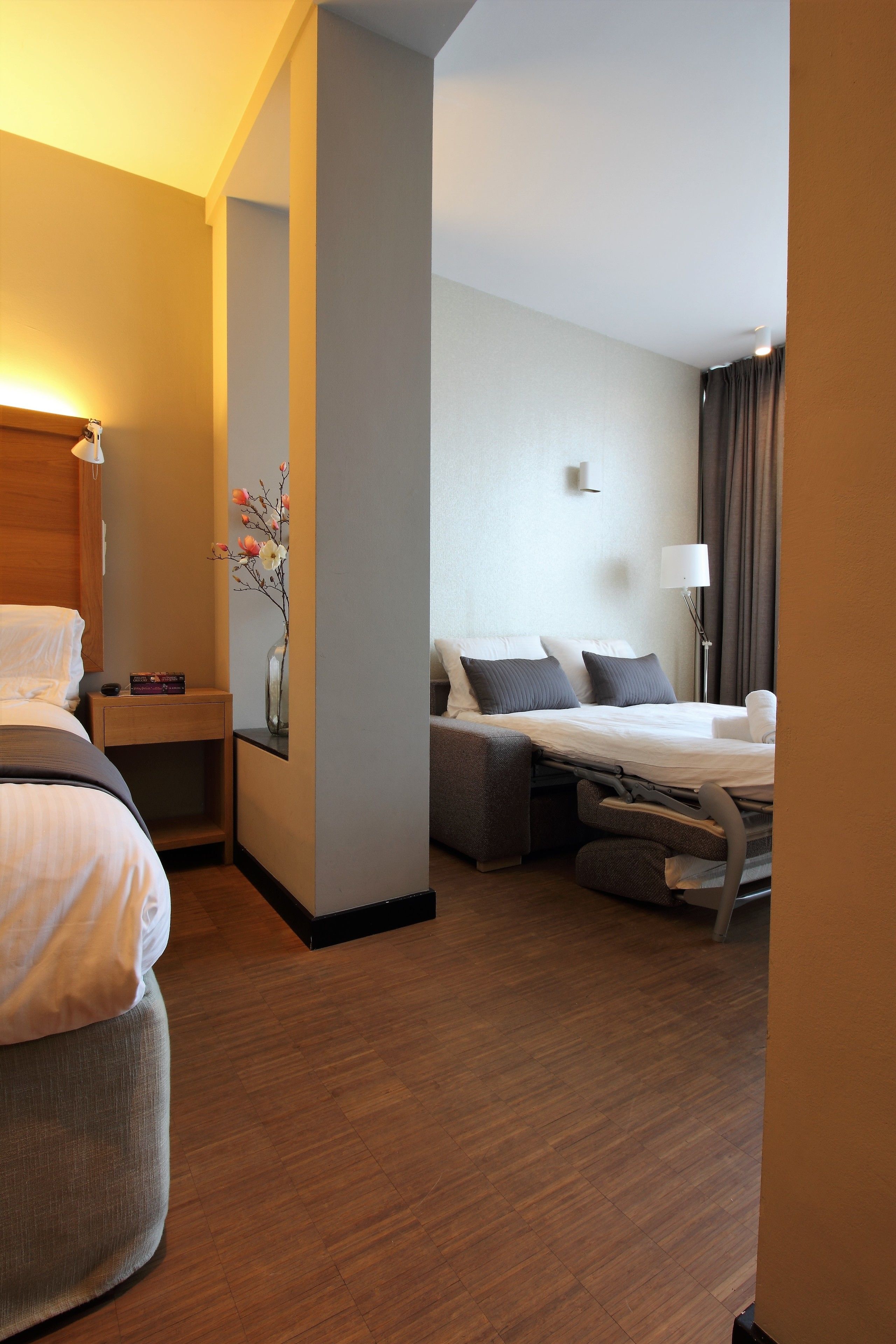 A hotel room with two beds, a bedside table with a lamp, a vase with flowers, and a large window with curtains.