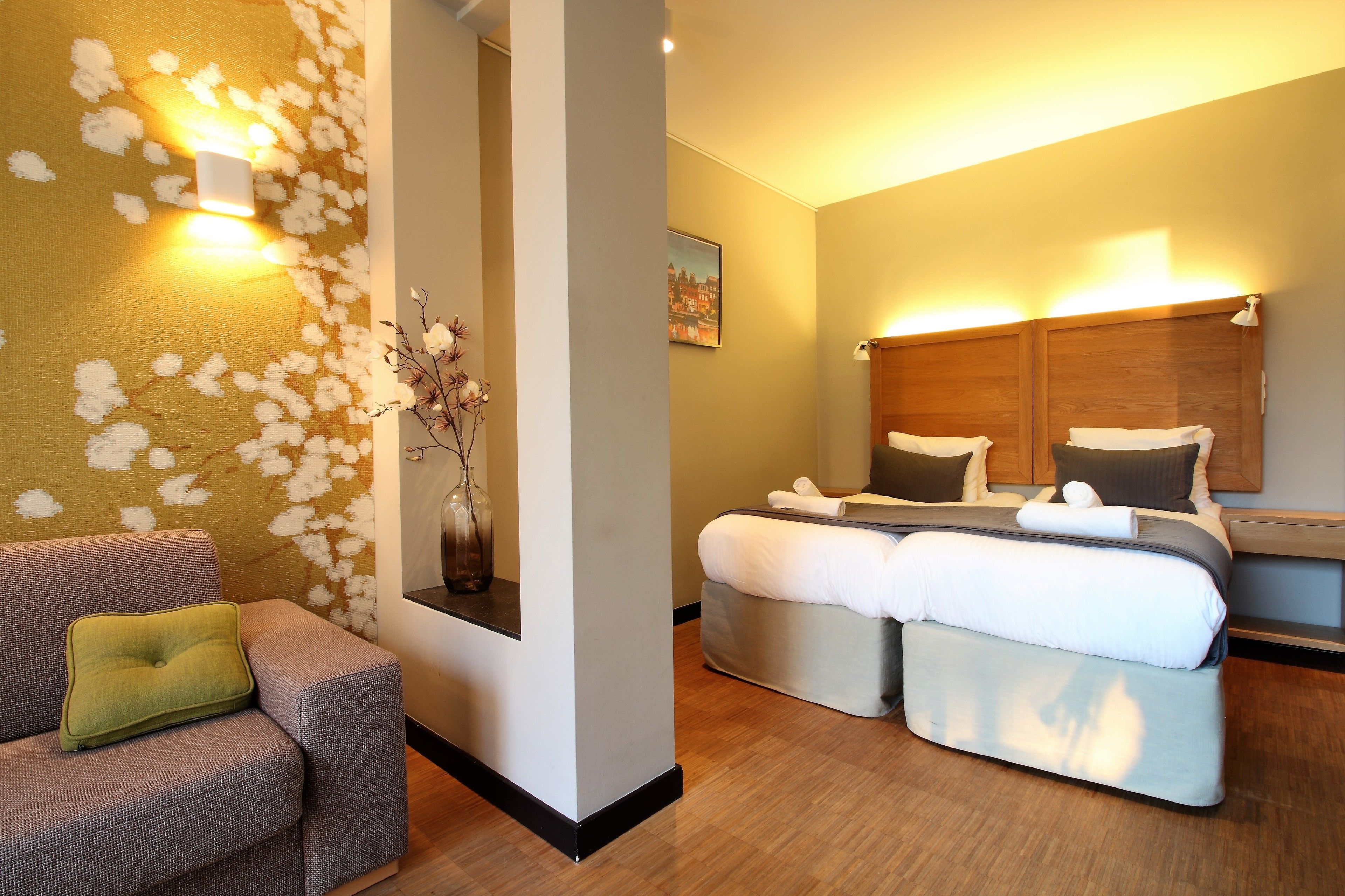 A hotel room with a sofa, a bed with two pillows, a nightstand, and a wall-mounted light fixture.