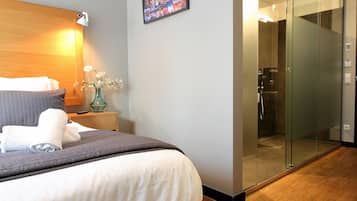 Habitación de hotel moderna con una cama grande, una mesita de noche con lámpara, un florero con flores, un box de ducha de vidrio y un cuadro de un paisaje urbano en la pared.