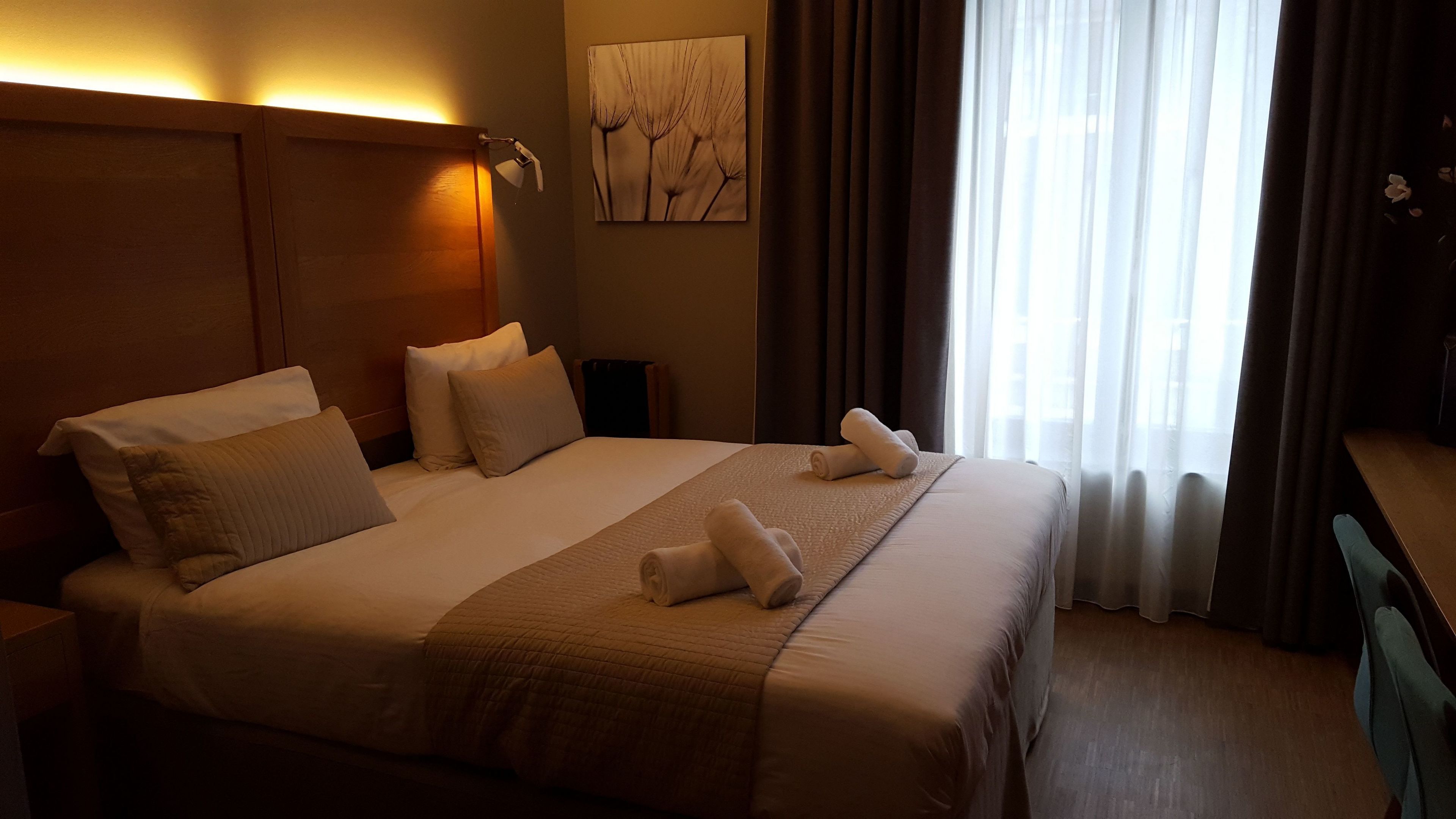 A hotel room with a large bed, two bedside tables, a nightstand, a wall-mounted lamp, a painting, and a window with curtains.