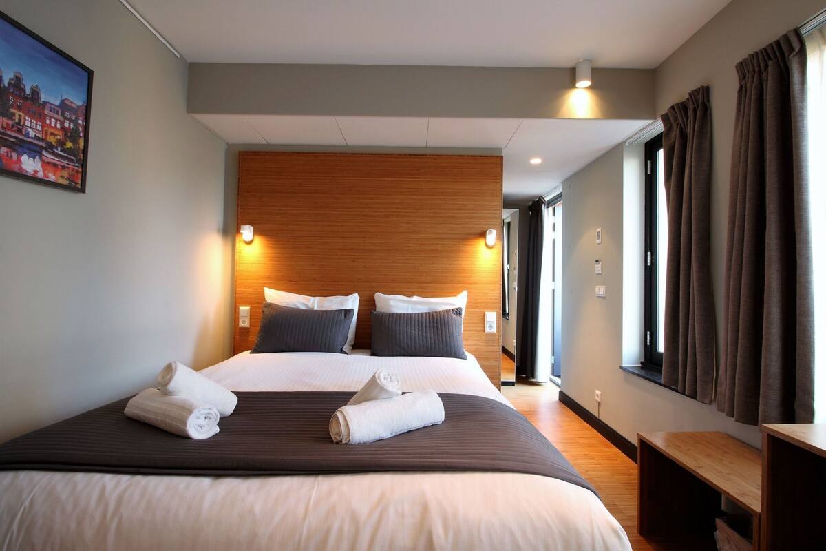 A hotel room with a large bed, wooden headboard, two bedside tables, a window with curtains, and a painting on the wall.