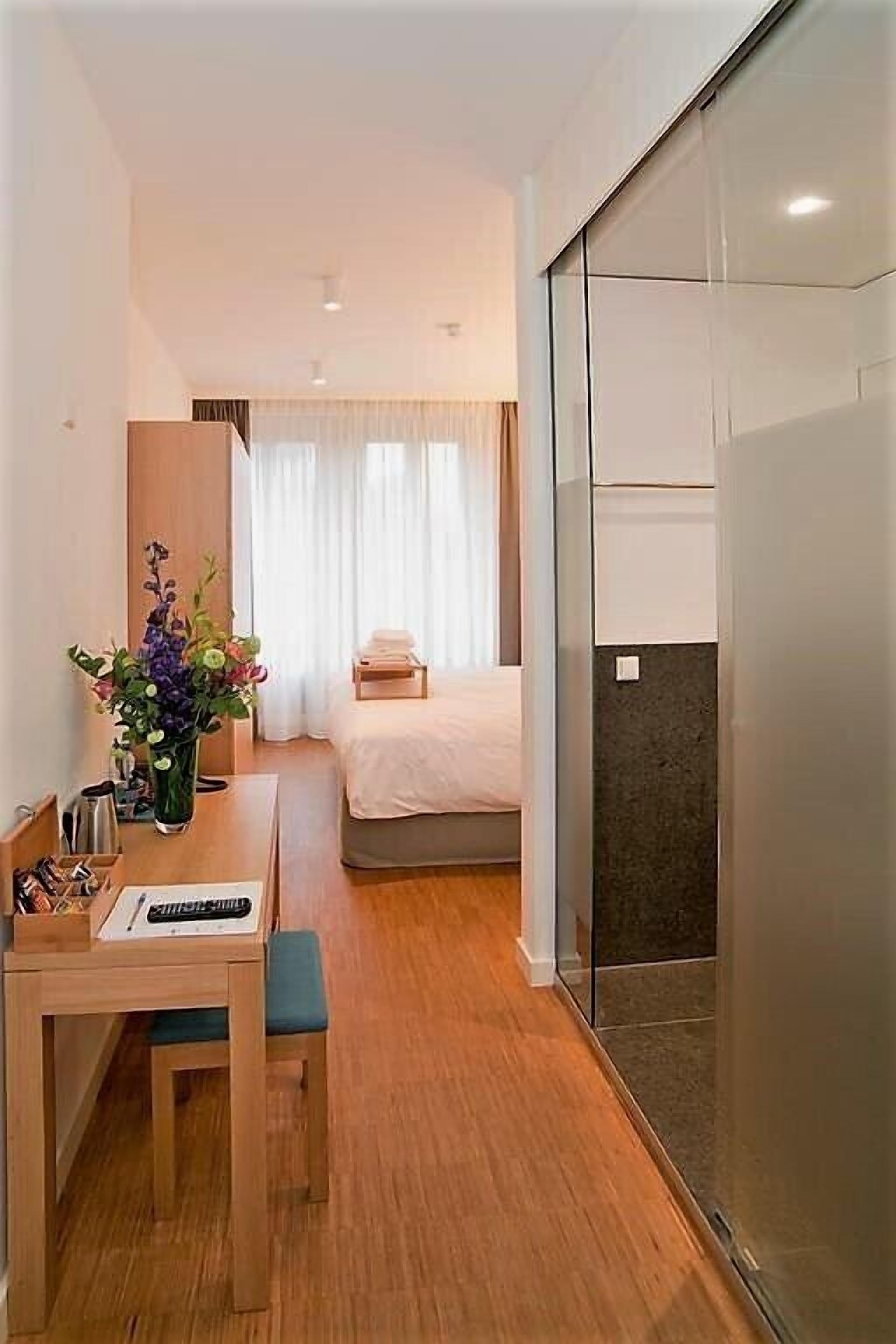 A hotel room with a bed, a desk with a chair, a vase of flowers, and a sliding glass door.