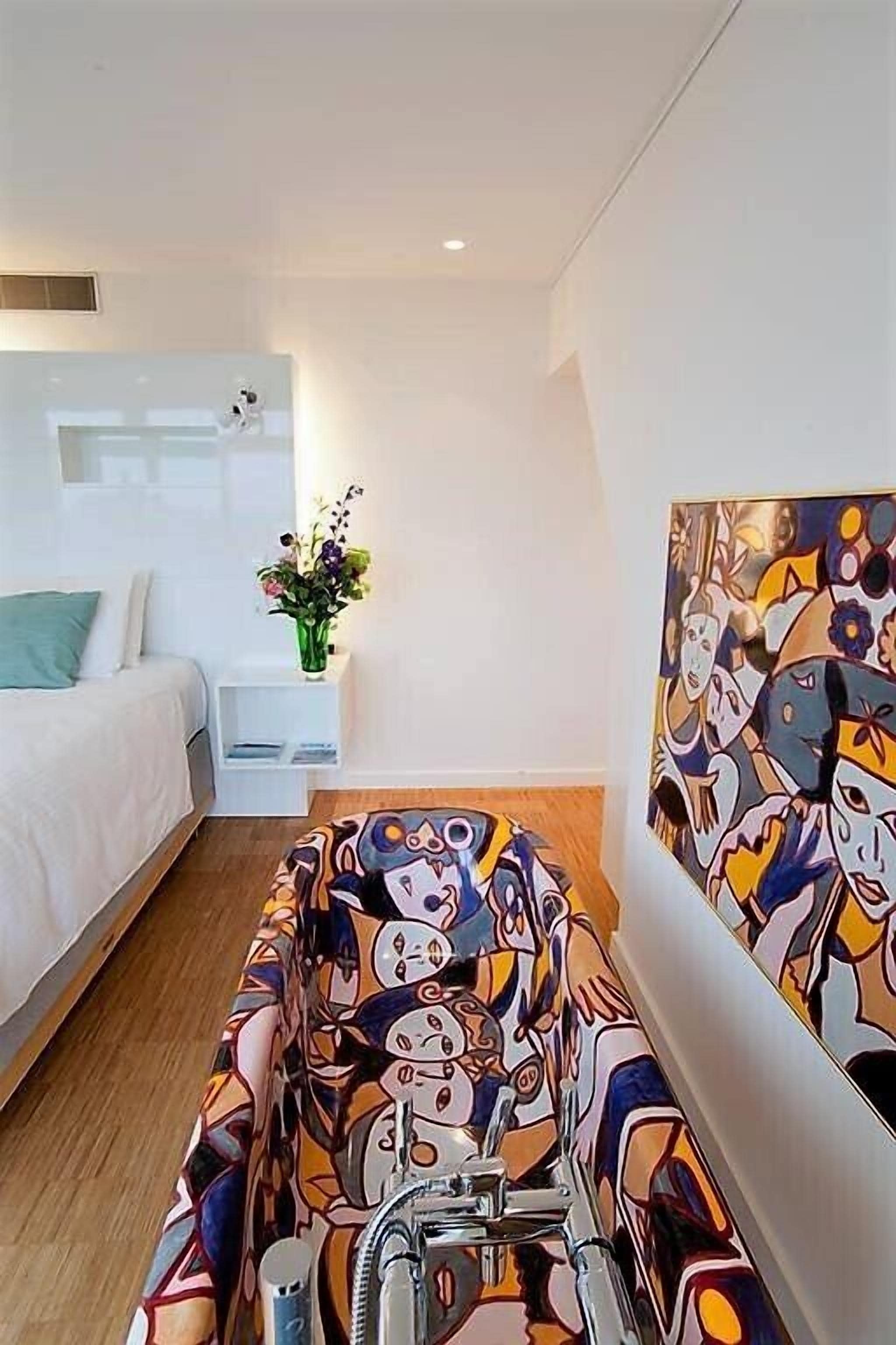 A modern bedroom with a bed, a bedside table with a vase of flowers, and a colorful abstract painting on the wall.