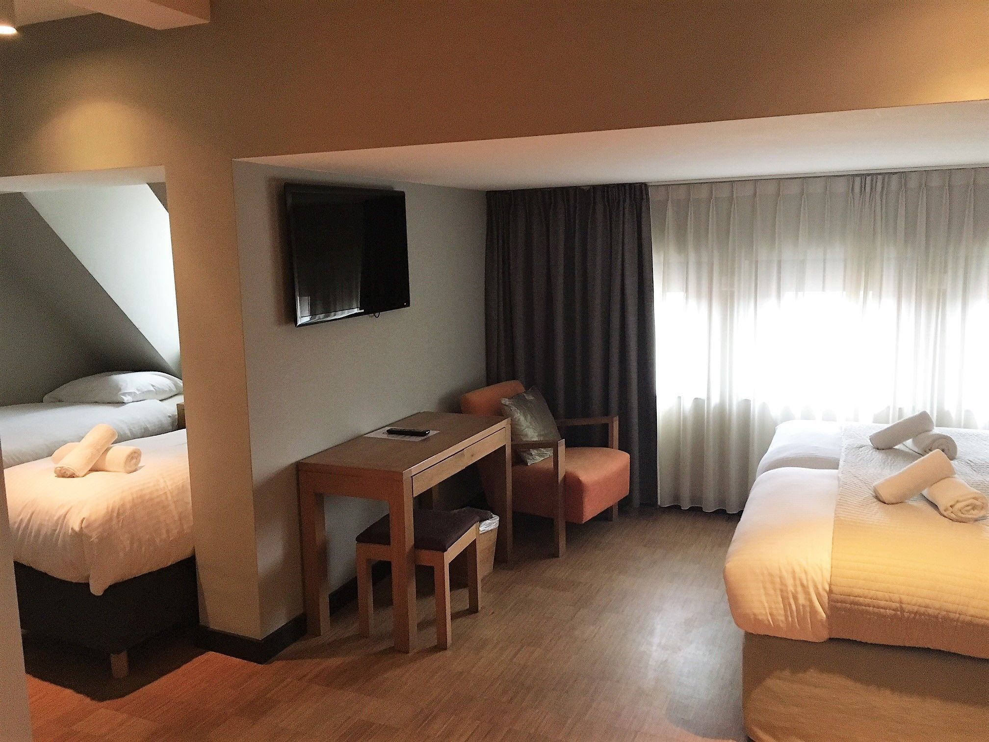 A hotel room with two beds, a desk, a chair, a TV, and a window with curtains.