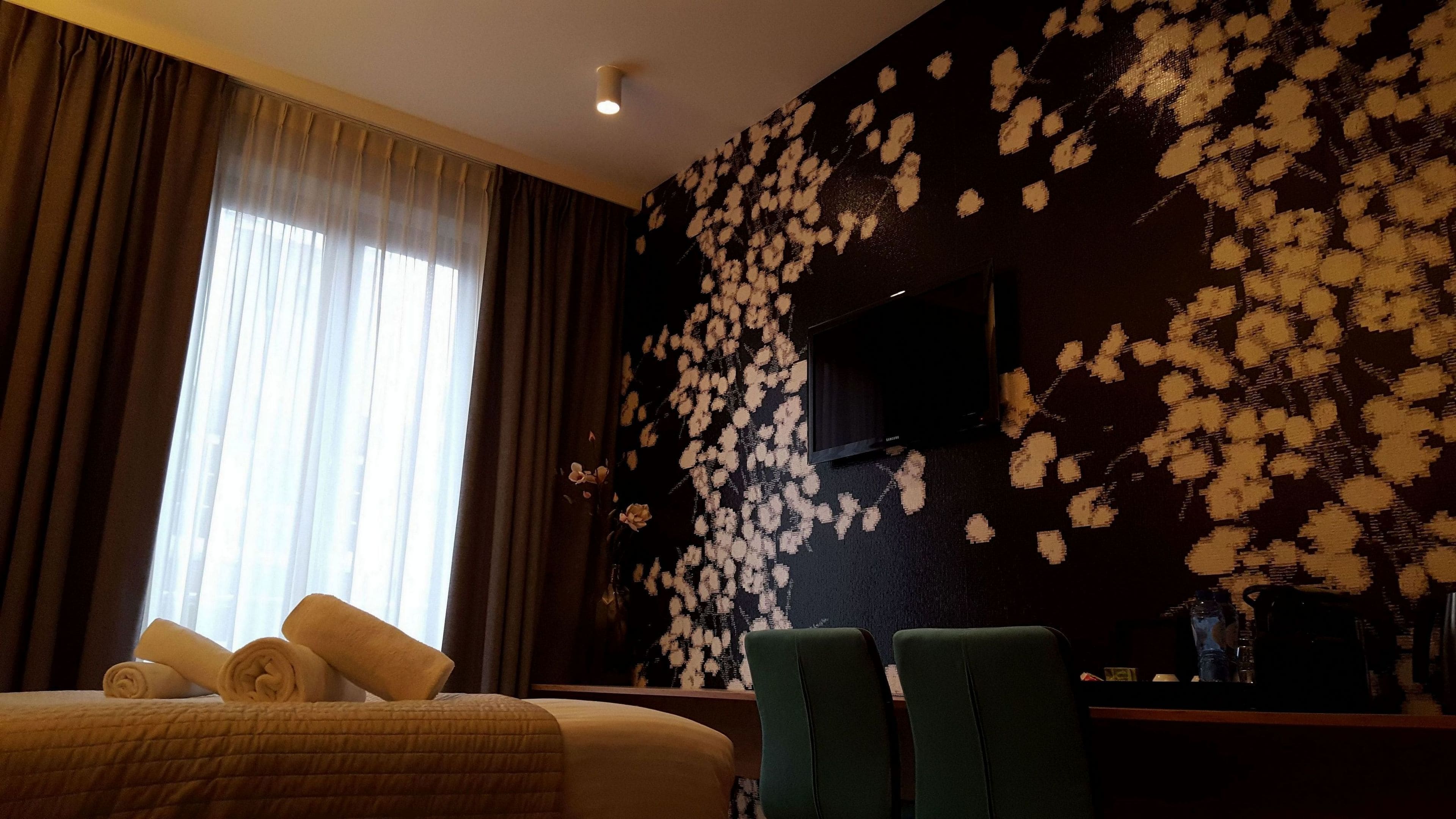 A hotel room with a bed, a desk, and a chair. There is a large window with curtains, and a wall with a floral pattern.