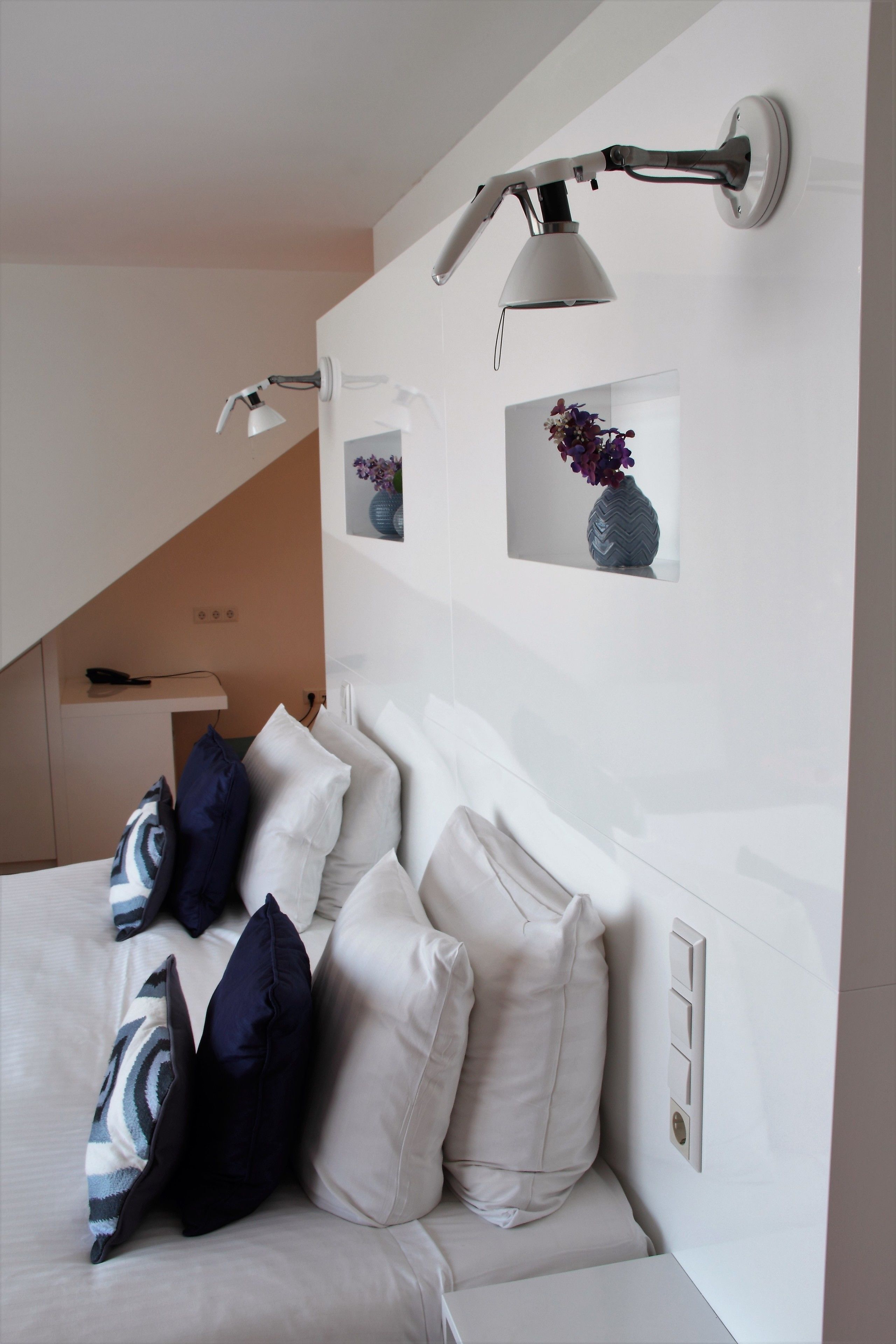 A modern bedroom with a white headboard, multiple pillows, a wall-mounted lamp, and a small framed picture of a vase.