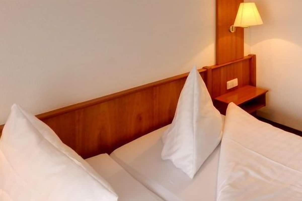 Habitación de hotel con cabecera de madera, ropa de cama blanca y mesita de noche con lámpara.