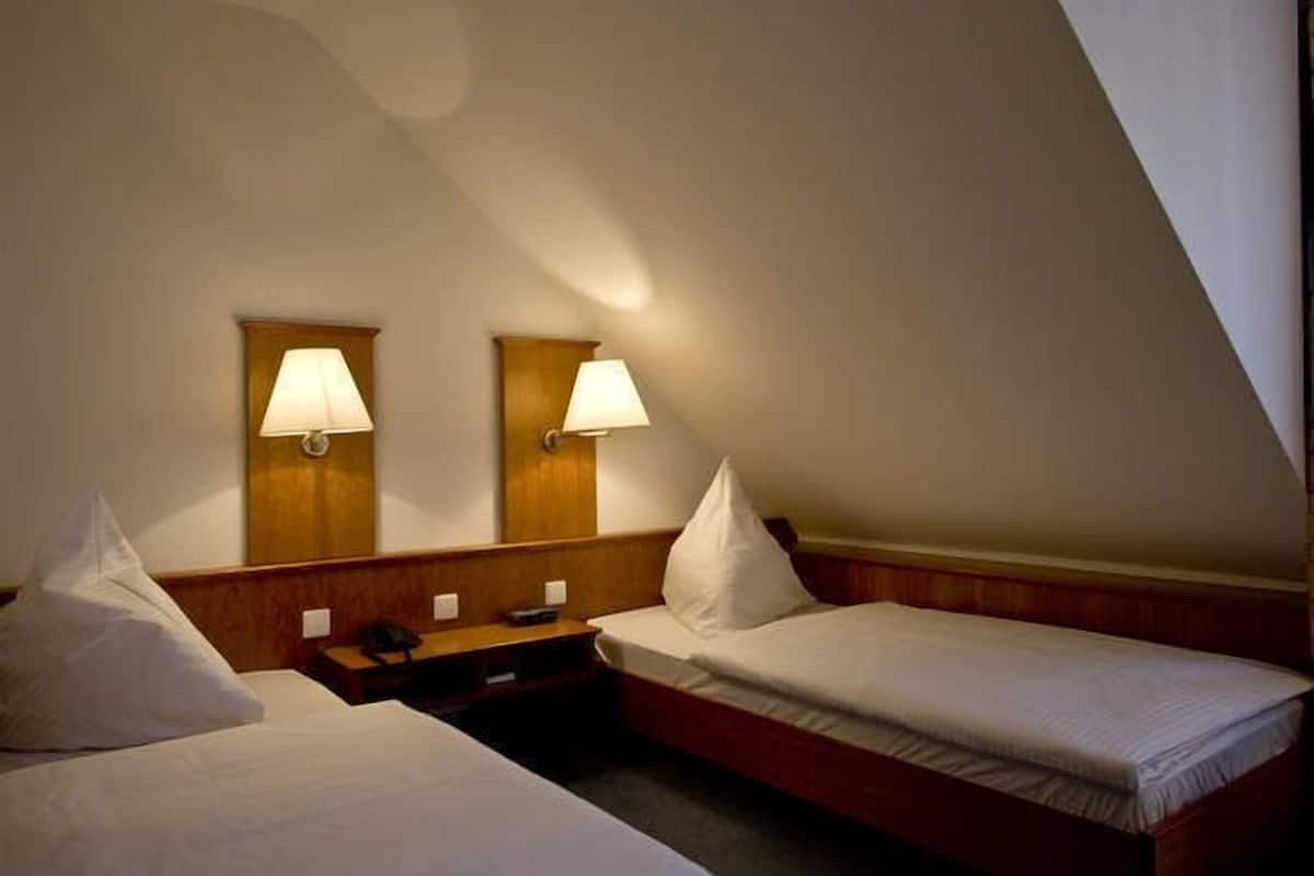 Habitación de hotel con dos camas individuales, cabecero de madera, mesitas de noche con lámparas y techo inclinado.