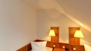 Quarto de hotel pequeno e individual, com cabeceira de madeira, duas mesinhas de cabeceira, uma pequena escrivaninha e uma luminária em cada lado.