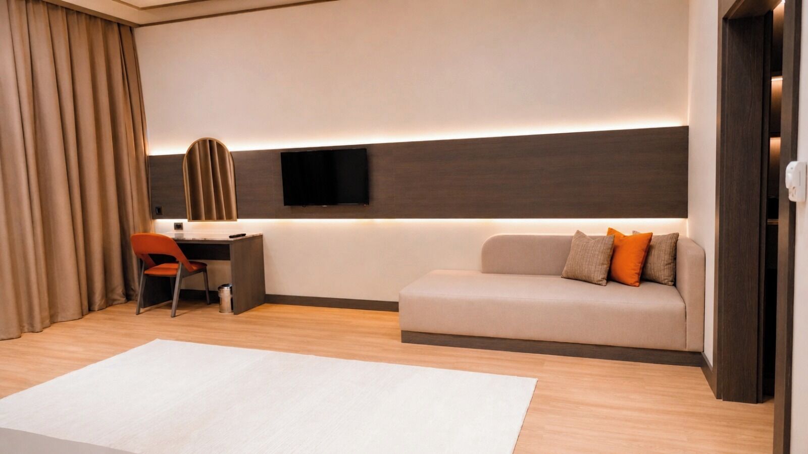 A modern hotel room with a desk, a sofa, a TV mounted on the wall, and a large window.