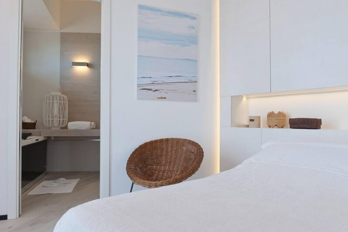 A modern bedroom with a large bed, a wicker chair, a wall-mounted shelf with decorative items, and a painting of a beach scene.