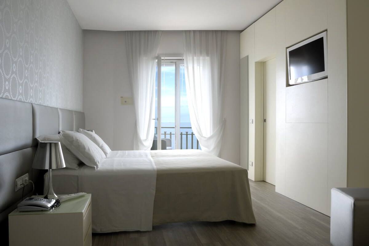A hotel room with a bed, a nightstand with a lamp, a TV mounted on the wall, and a view of the sky through sheer curtains.