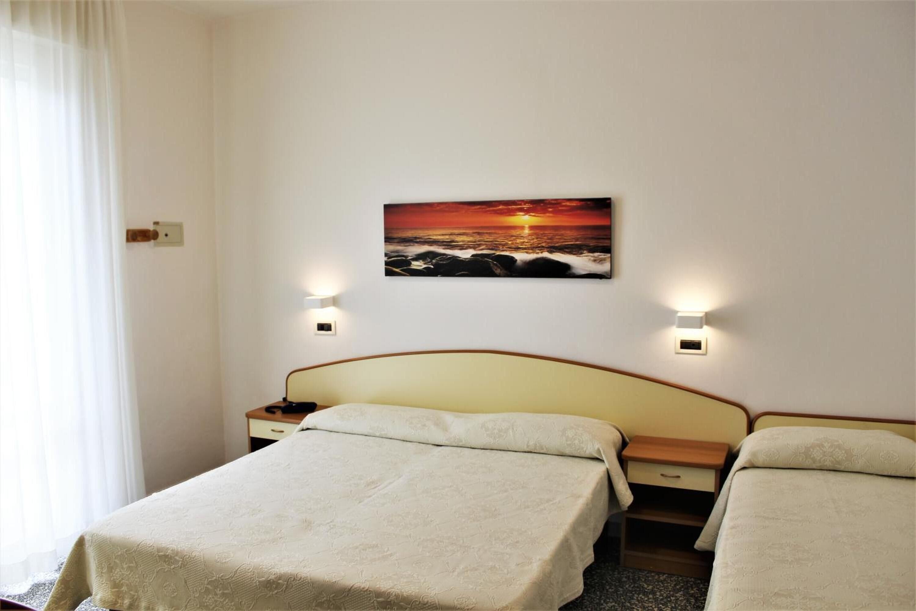 A hotel room with two beds, a painting of a sunset over the ocean on the wall, and bedside lamps.