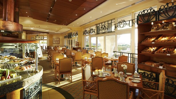 Ein Speisesaal eines Hotels mit Korbstühlen, einem Buffet mit verschiedenen Speisen und Blick ins Freie durch große Fenster.