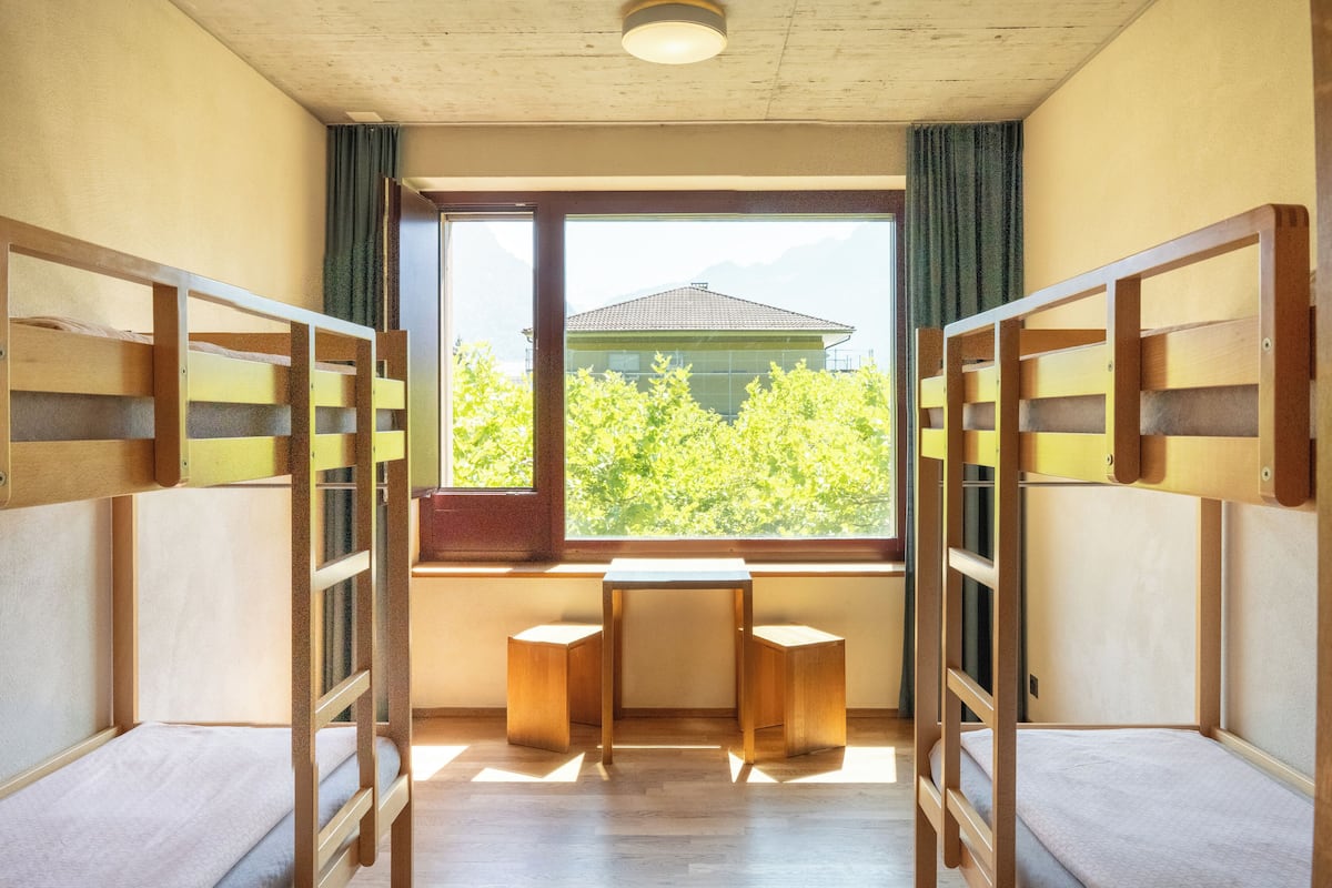 A room with bunk beds, a small table, and a window with a view of trees and a building.