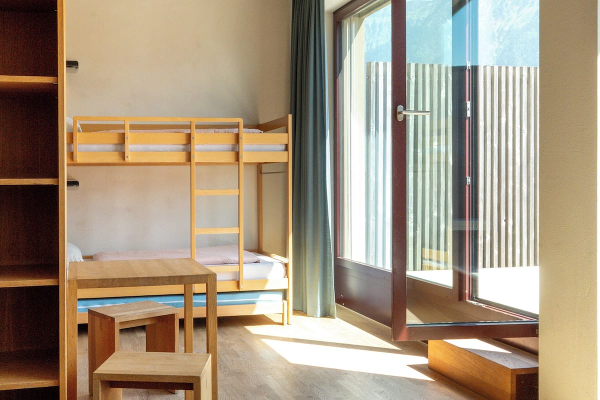 A room with bunk beds, a wooden desk, and a large window with a view of mountains.