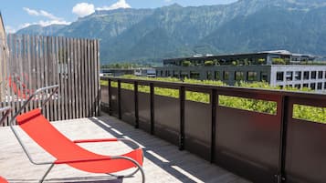 A balcony with red chairs, a wooden fence, and a view of mountains and a building.