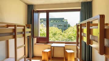 Quarto com beliches, uma mesa pequena e janela com vista para edifícios e árvores.