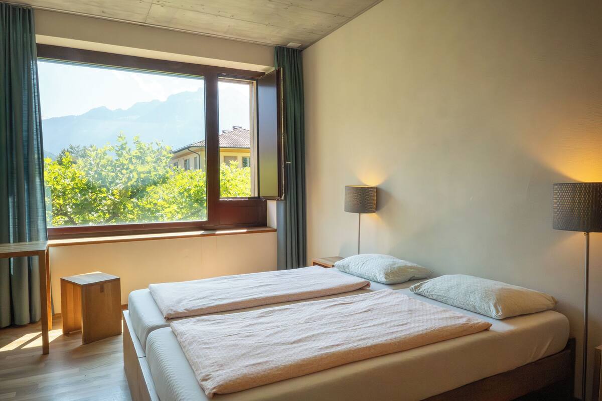 A bedroom with a large window, a bed, a bedside table, and a lamp.