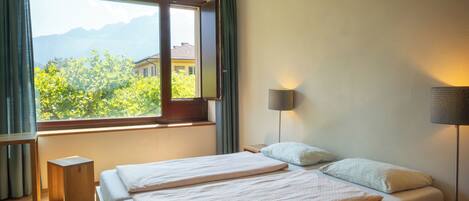 A bedroom with a large window, a bed, a bedside table, and a lamp.