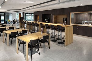 Una cafetería moderna con largas mesas de madera y sillas negras, una máquina de café y un amplio ventanal que proporciona luz natural.