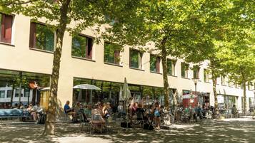 An outdoor café with tables and chairs, a building with large windows, and trees providing shade.